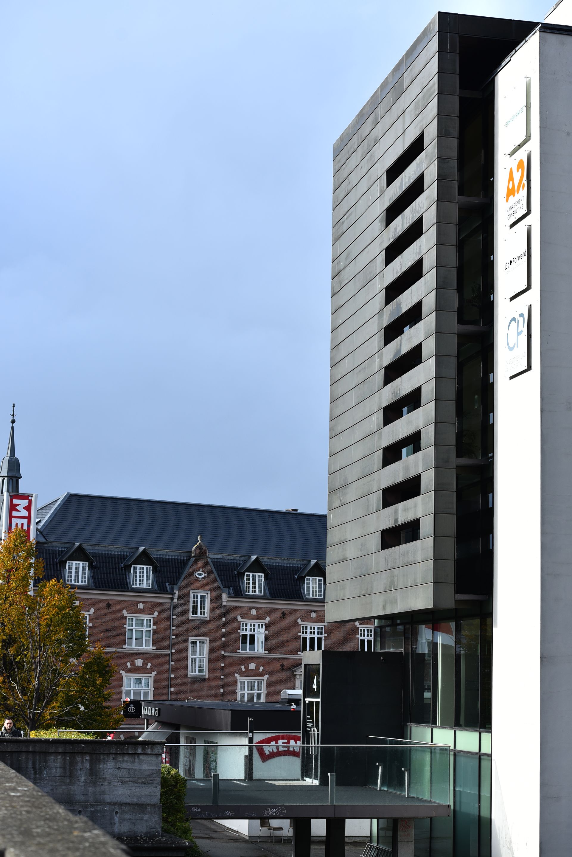 Modern svart byggnad med fönsterfasad bredvid en vit byggnad. Röd tegelbyggnad i bakgrunden. Molnig himmel.