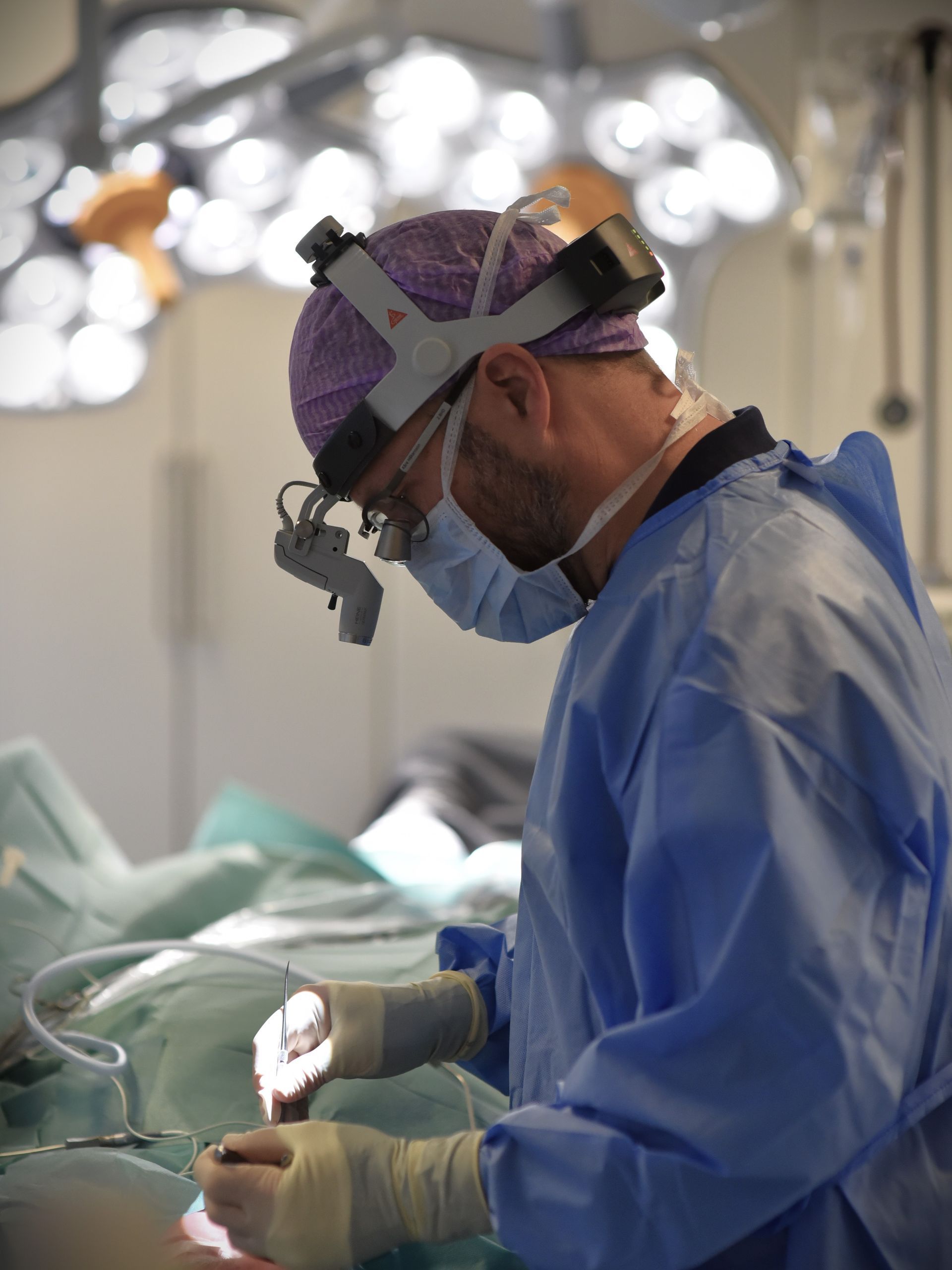 Kirurg i blå scrubs opererar under starkt ljus, med pannlampa och mask.