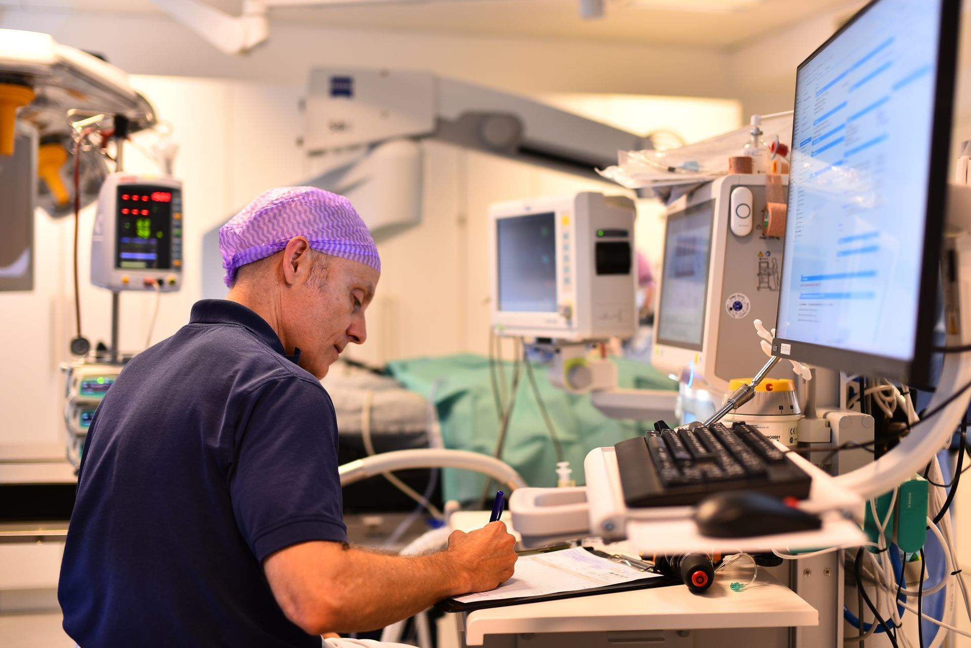 En person med operationsmössa skriver på ett urklipp i en operationssal, bredvid skärmar och utrustning.