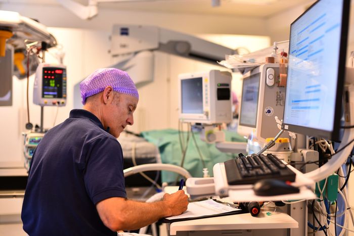 En person med operationsmössa skriver på ett urklipp i en operationssal, bredvid skärmar och utrustning.
