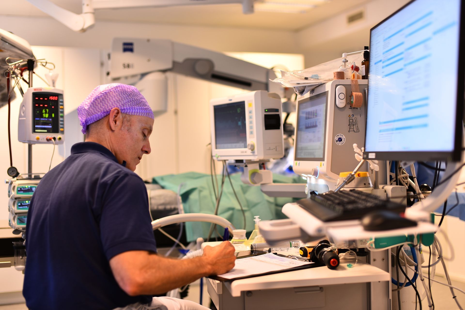 Kirurg i operationsmössa skriver vid ett skrivbord, omgiven av medicinska monitorer och utrustning i en operationssal.