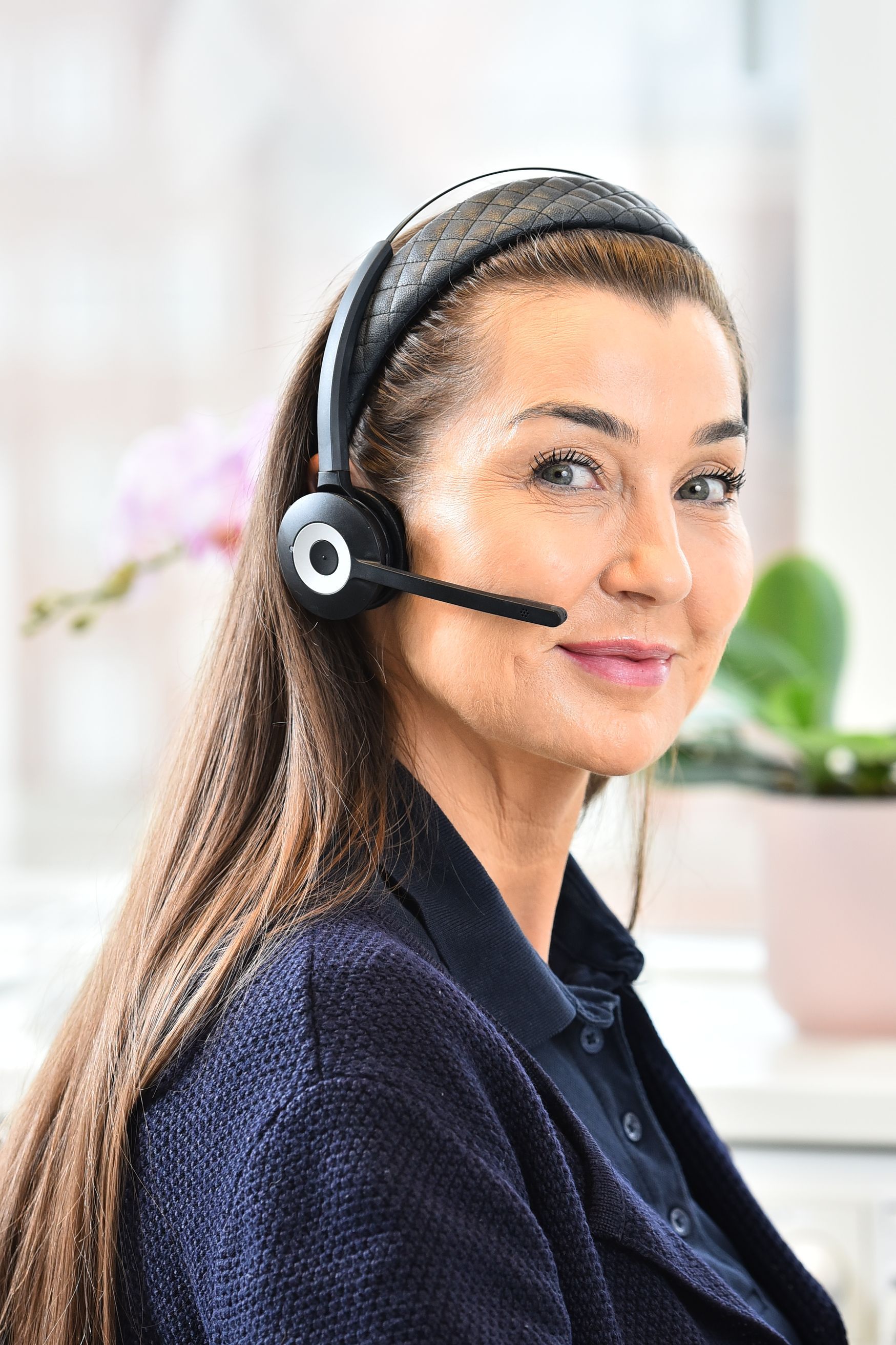 Kvinna med headset, leende, med långt brunt hår, nära fönster och krukväxt.