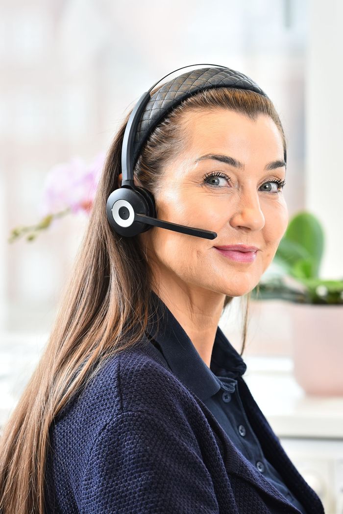 Kvinna med headset, leende, med långt brunt hår, nära fönster och krukväxt.