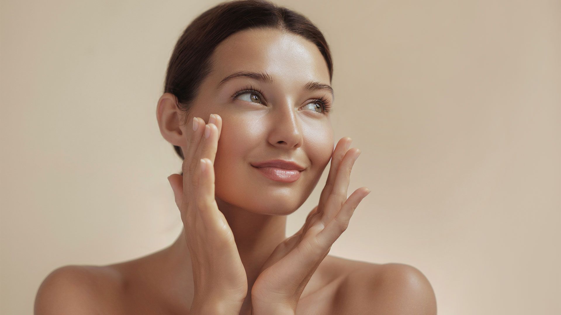Woman with hands near face, gazing upward, against a neutral background; radiant skin.
