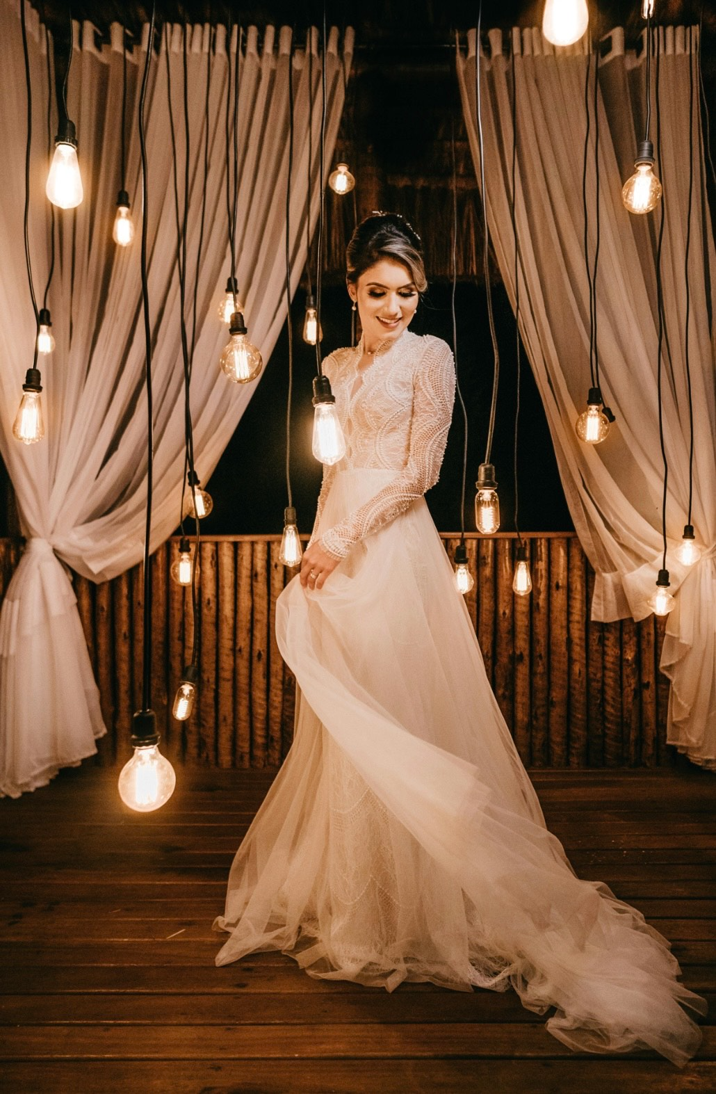 Bride in white dress poses on a wooden deck lit by hanging lights.