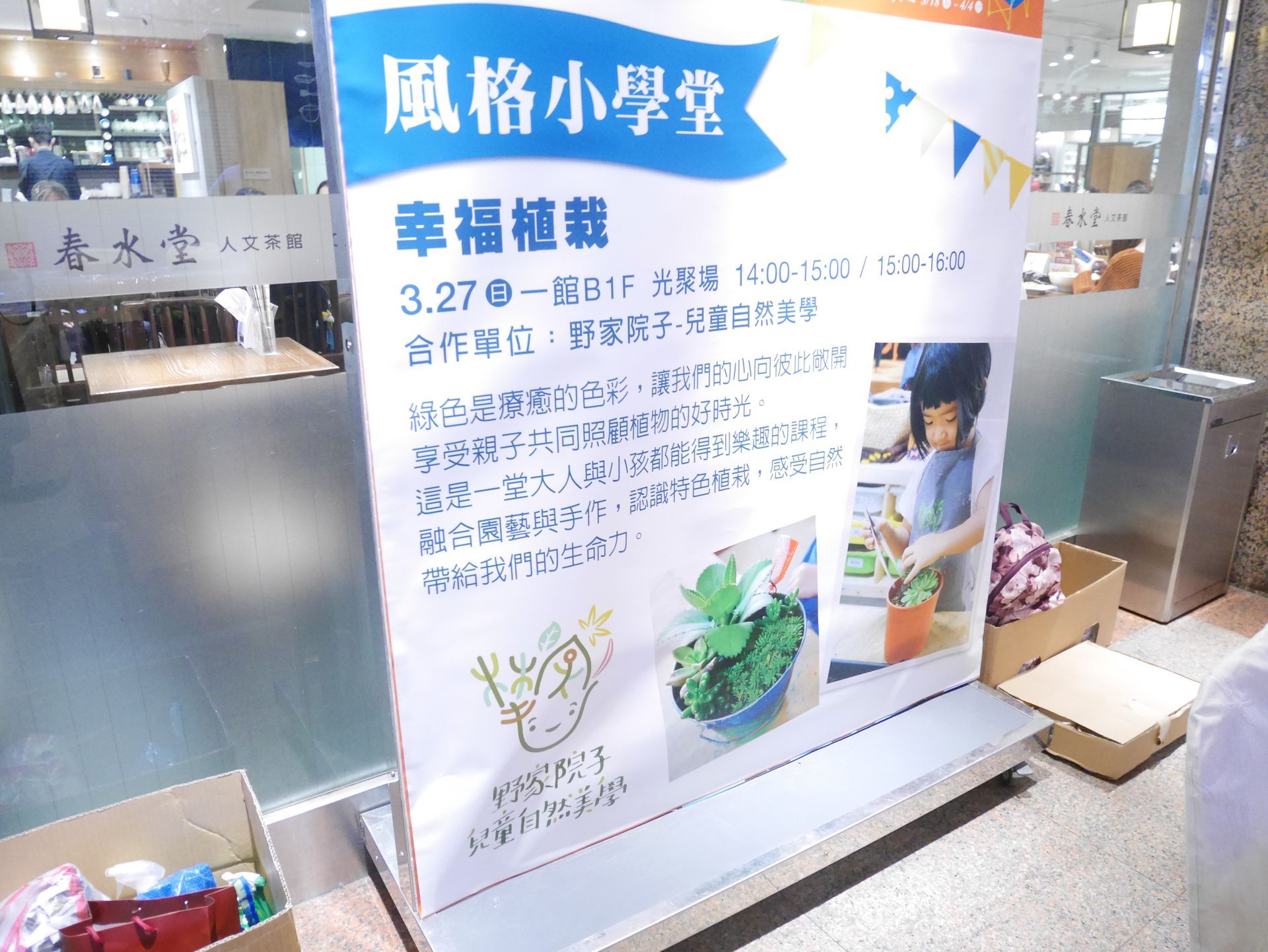 購物中心內的大型看板，上面有中文文字和照片，宣傳以春天為主題的校園活動。