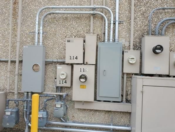 Electrical boxes and conduits on a building exterior. Several gray and tan boxes, with meters, are mounted to a textured wall.
