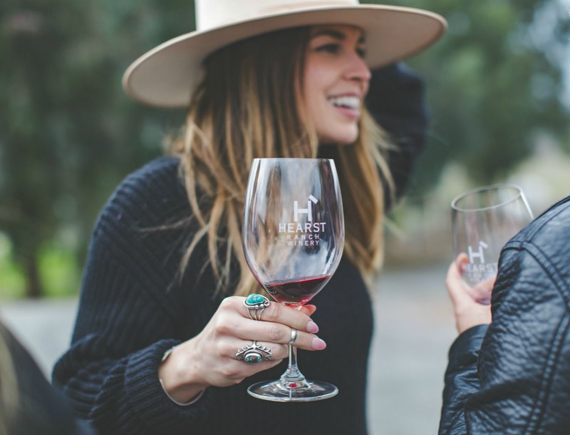 Woman in a wide-brim hat holding a wine glass outdoors, smiling and chatting with another person