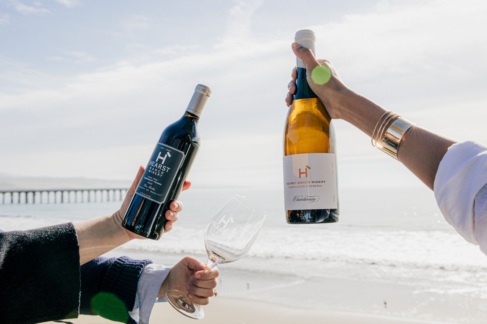 Two hands hold champagne bottles and glasses at a sunny beach celebration with a pier in the background