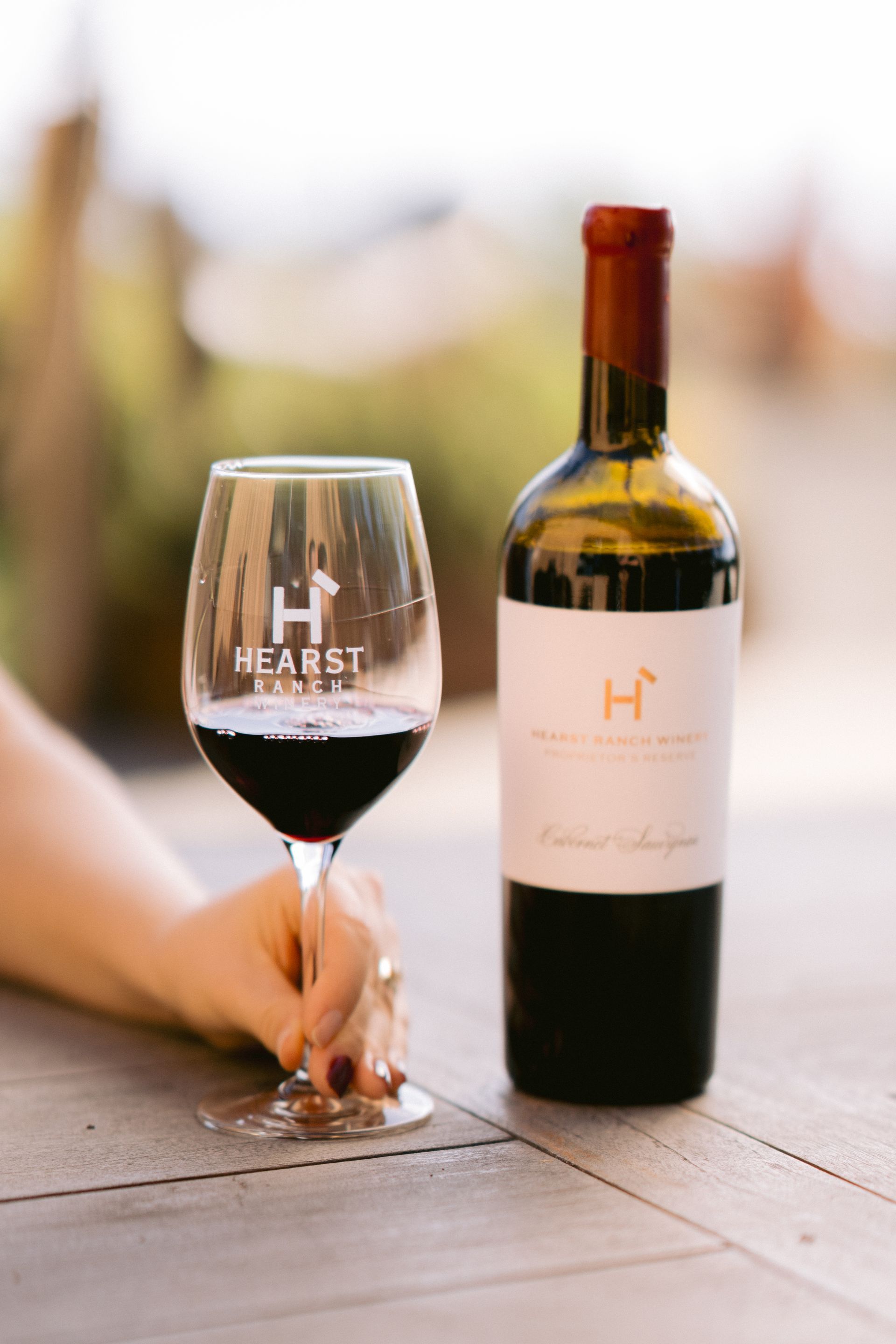 Wine glass and bottle on a table, with a hand holding the glass in a softly blurred outdoor setting