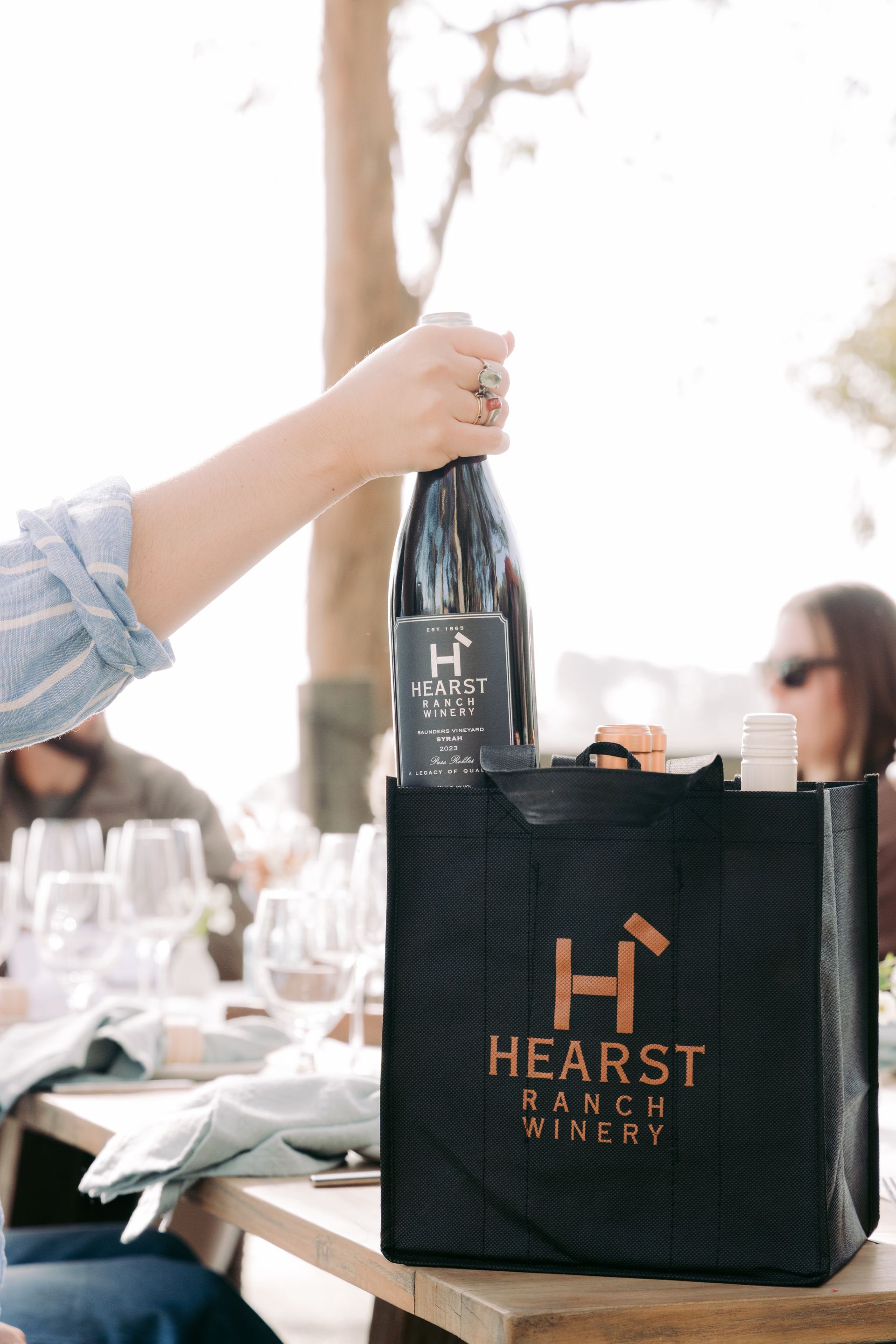 Hand placing a wine bottle into a black Hearst Ranch shopping bag on a table outdoors