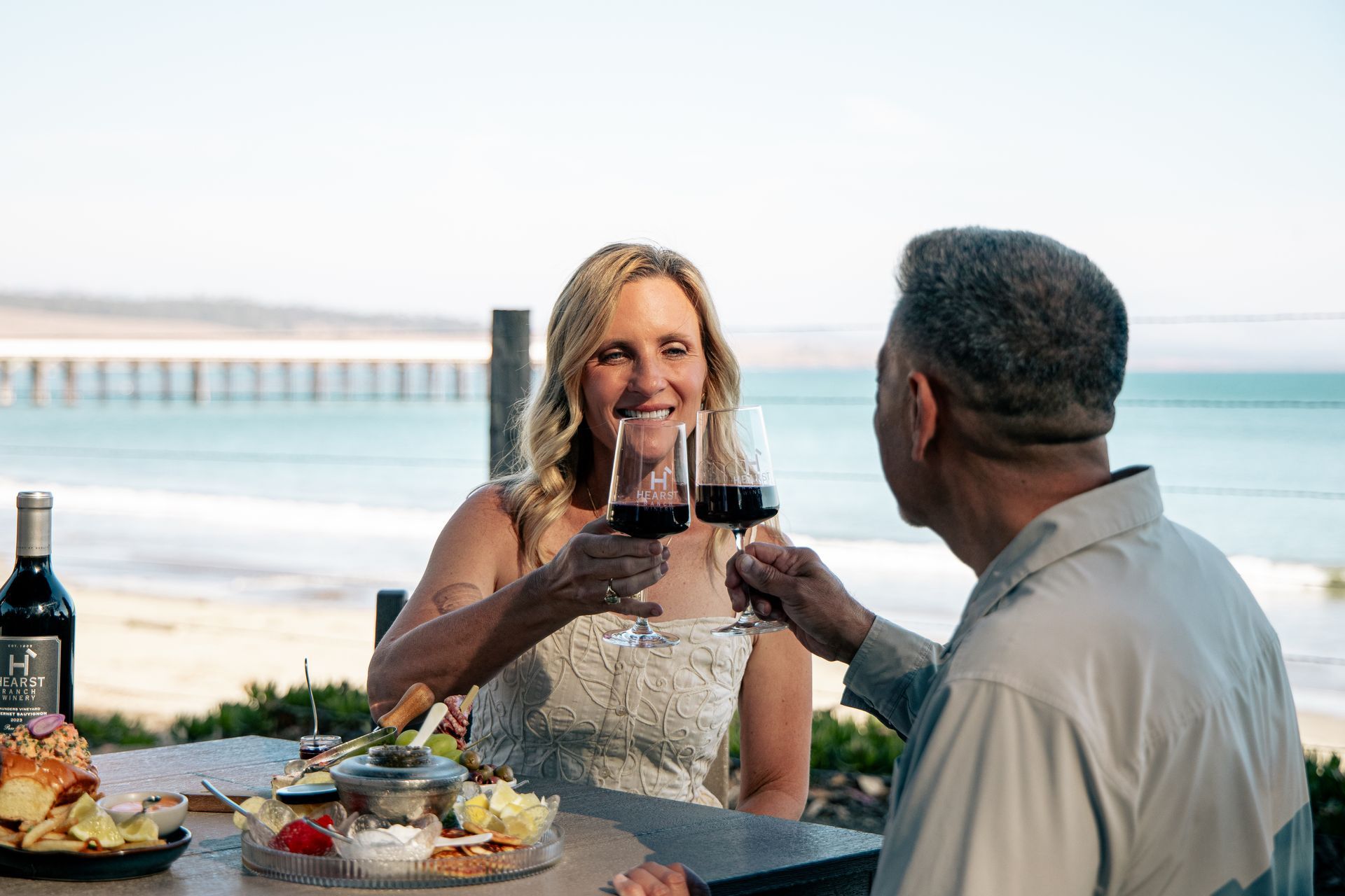 Two people clink red wine glasses by a beach with a pier in the background