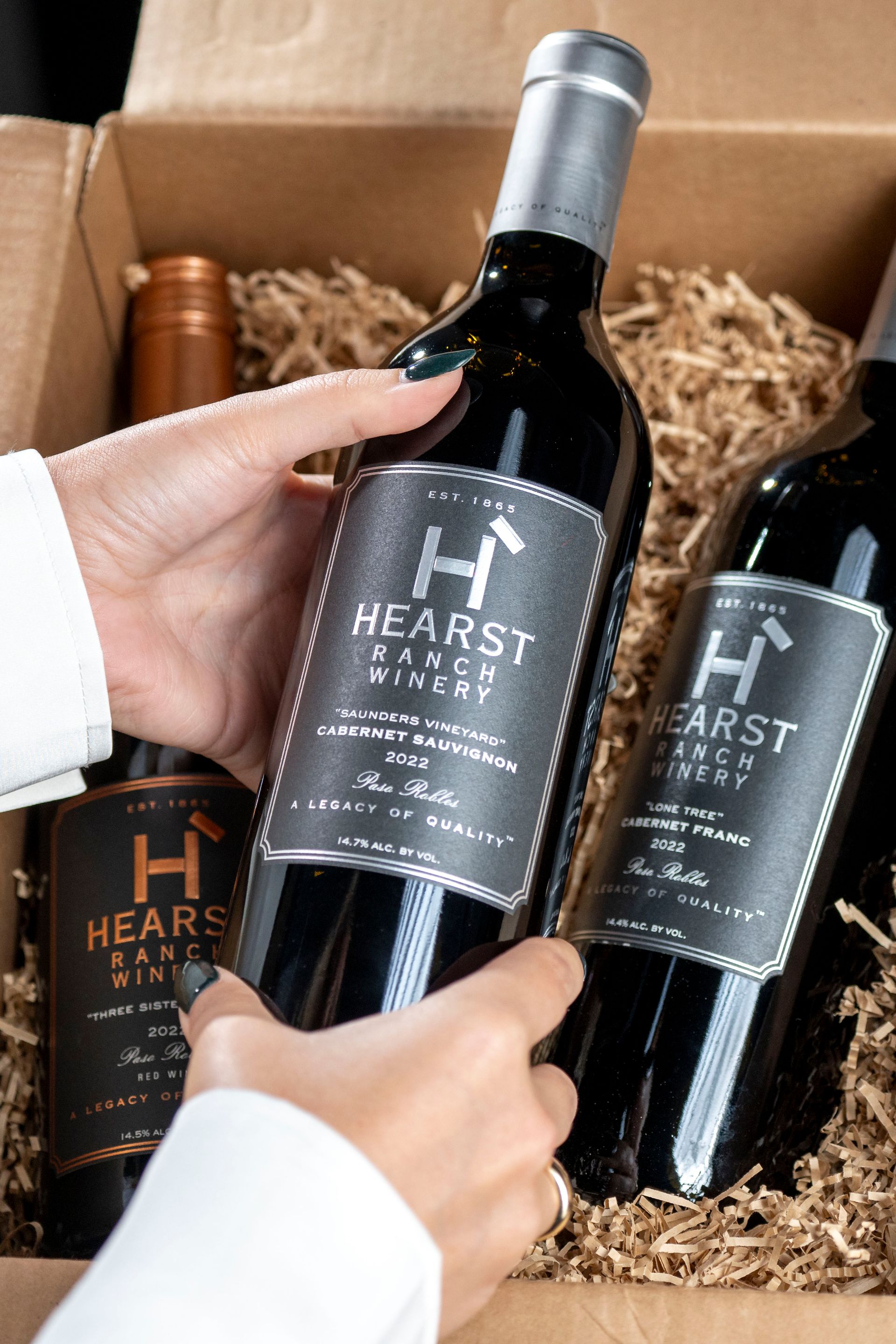 Hands holding a black Heart Winery wine bottle in a gift box with two other bottles and shredded packing paper