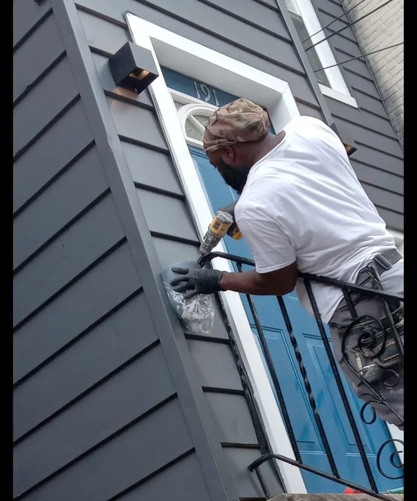 A person in a white shirt and head covering uses a power drill on a house exterior next to a blue door and black railing.