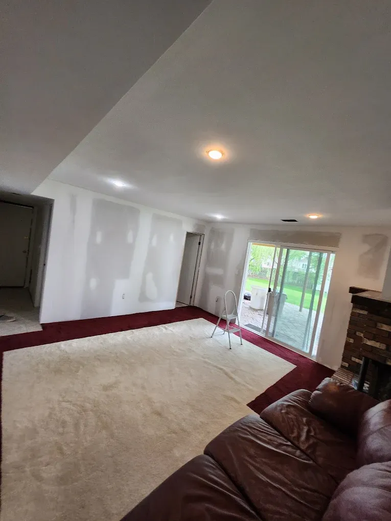 A room under renovation with drywall patches on walls, beige carpet, a red floor border, and a leather sofa.