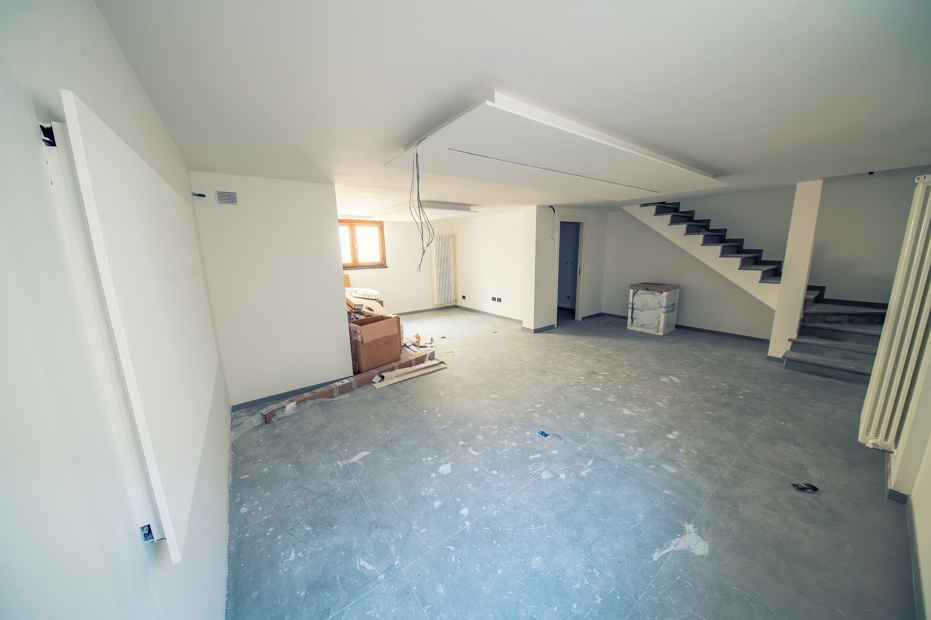 An unfinished room with gray flooring, white walls, a staircase, and a central ceiling light fixture.