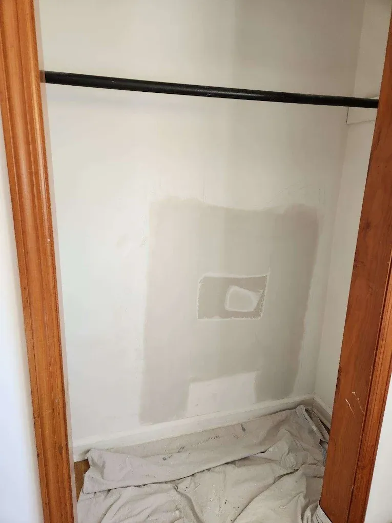 A closet interior shows a freshly patched drywall repair with a square light gray area on a white wall and a drop cloth.
