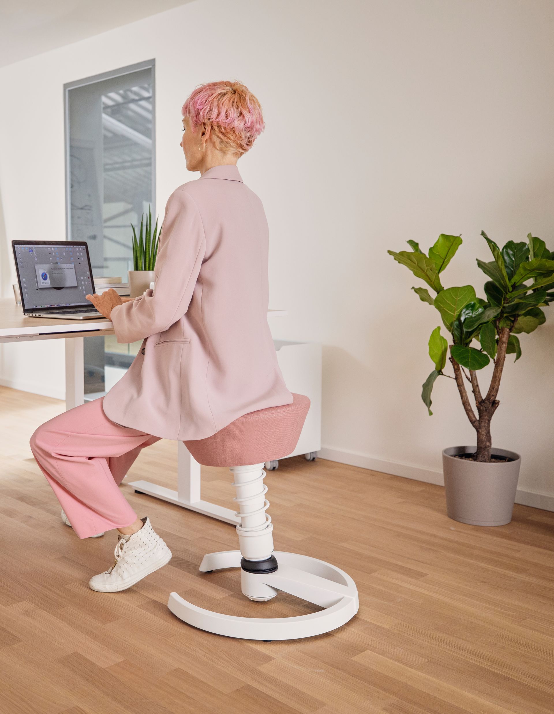 Frau arbeitet im Home Office auf ergonomischem Hocker vor Laptop und Pflanzen

