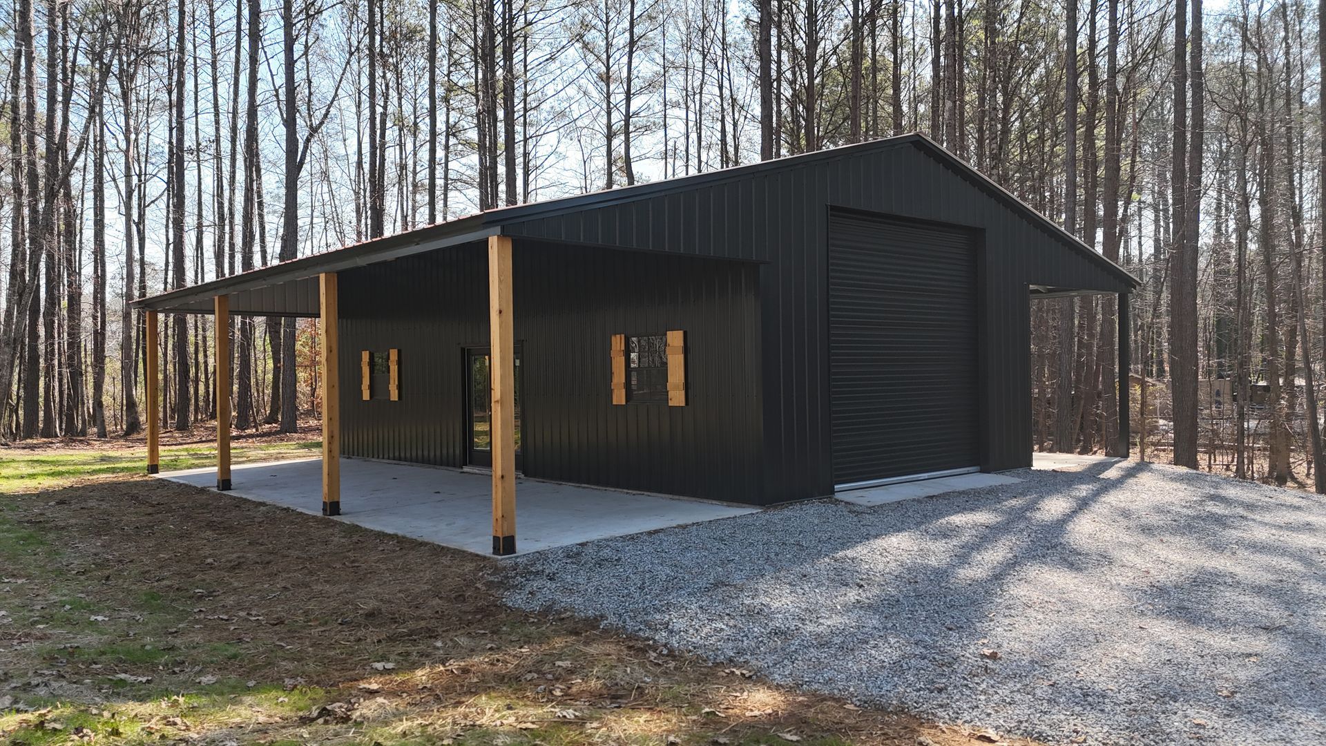 Black metal building with a carport on a concrete pad, gravel ground, and trees in the background.