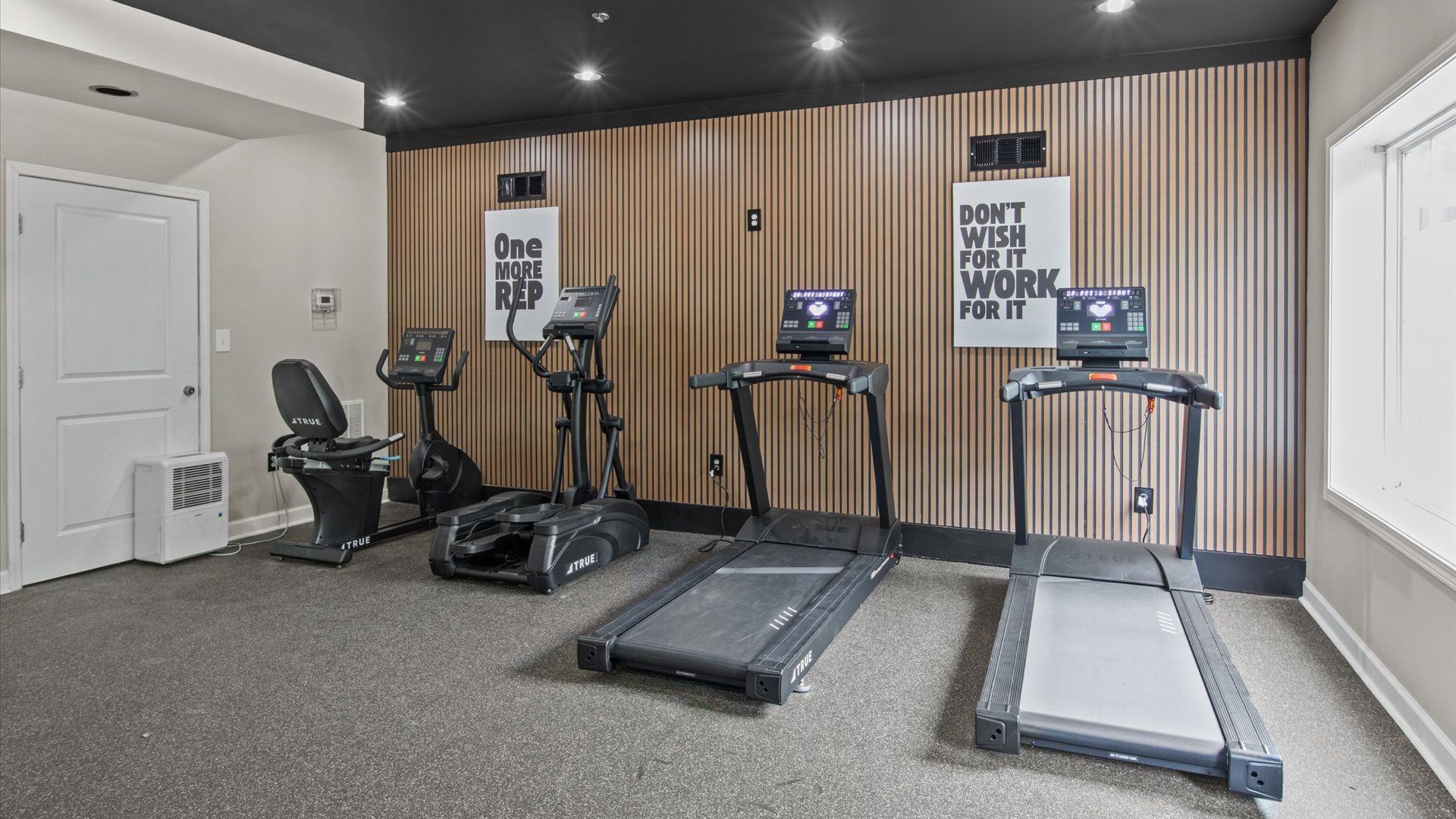 A fitness room with a treadmill, elliptical, and recumbent bike, featuring a large wall mirror and an exit door.