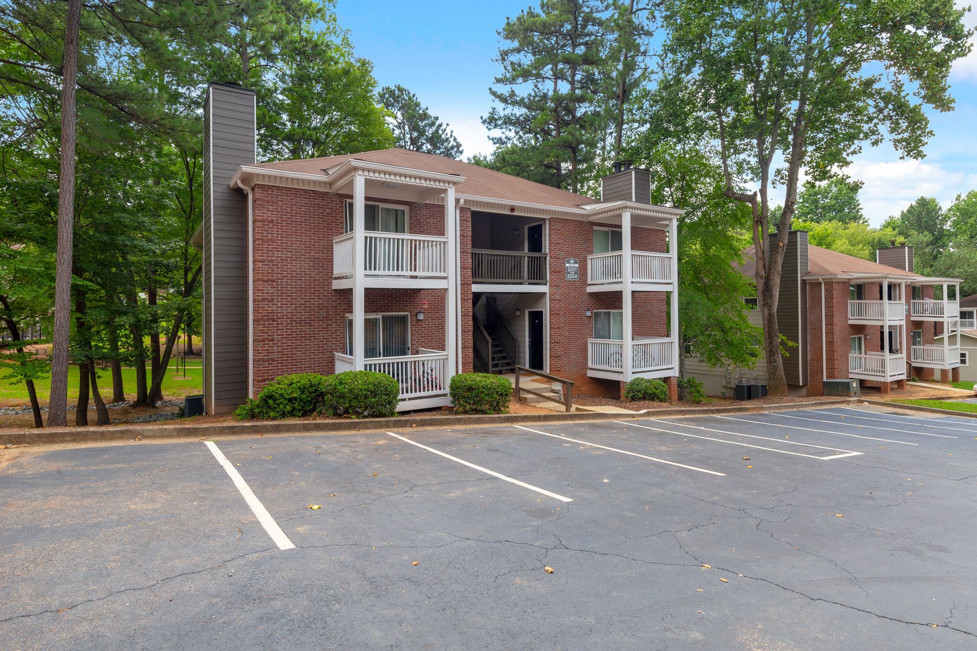 A two-story red brick apartment complex with white balconies, surrounded by trees next to an asphalt parking lot.