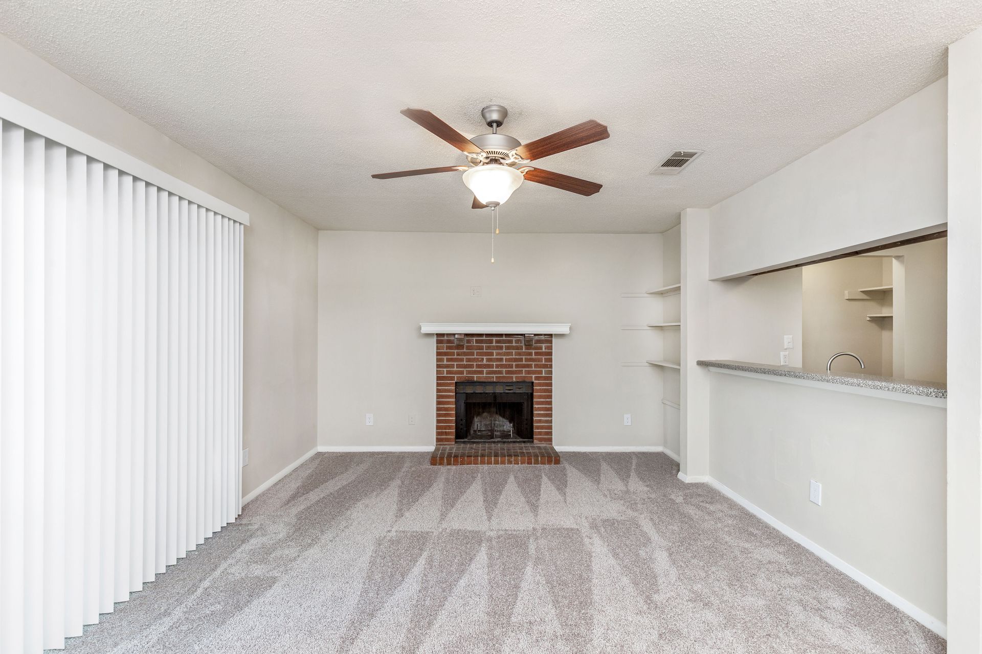 An empty living room with gray carpet, a brick fireplace, a ceiling fan, and an open bar counter to a kitchen area.