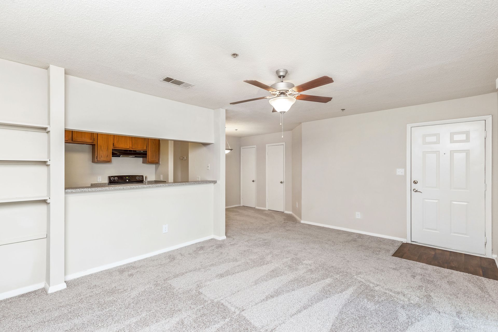 A bright, neutral-toned living room with grey carpet, a ceiling fan, a kitchen pass-through, and a white entry door.