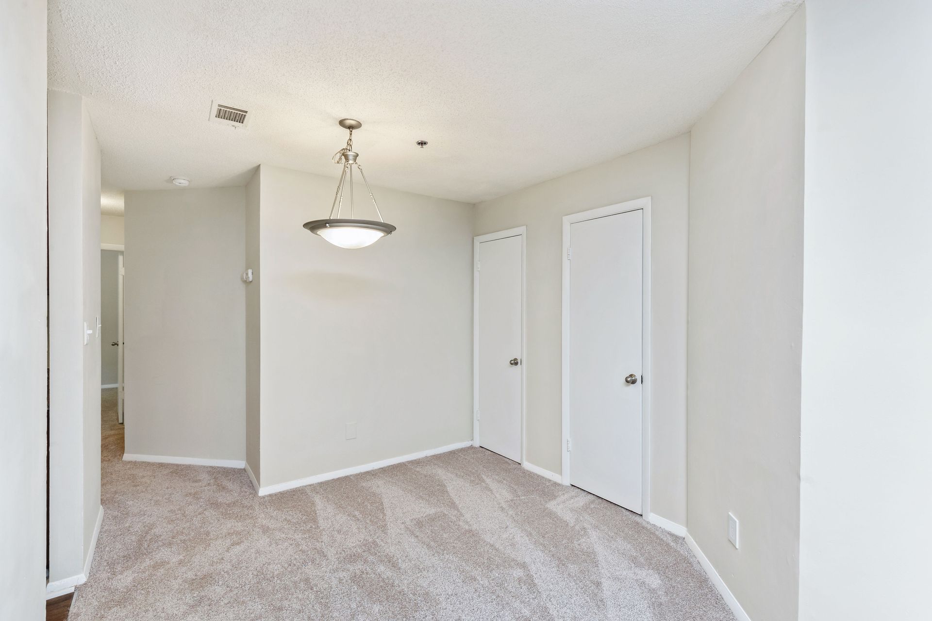 An empty carpeted dining area featuring a modern light fixture, plain walls, and two closed white doors.