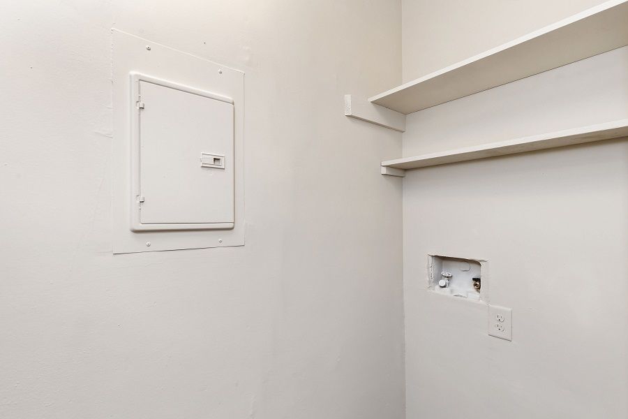 A white interior wall featuring a closed utility panel, two white floating shelves, and an exposed laundry hookup box.