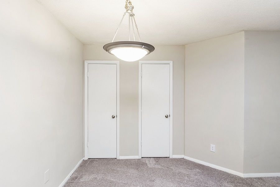 An empty room with light gray walls, gray carpet, two white doors, and a pendant light fixture hanging from the ceiling.