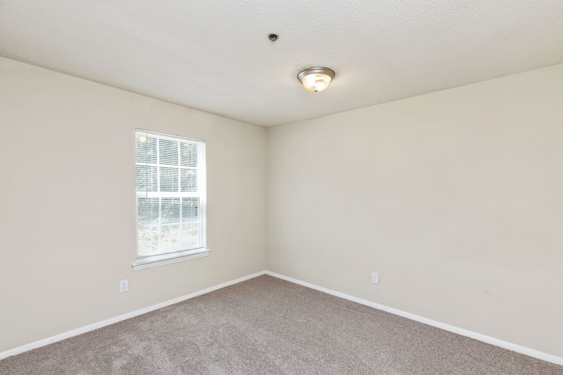 An empty bedroom with beige walls, gray carpeting, a single window with blinds, and a circular ceiling light fixture.