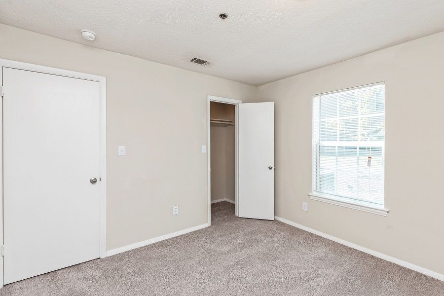 Empty bedroom with light beige walls, gray carpeting, a window, a closed door on the left, and an open closet on the right.