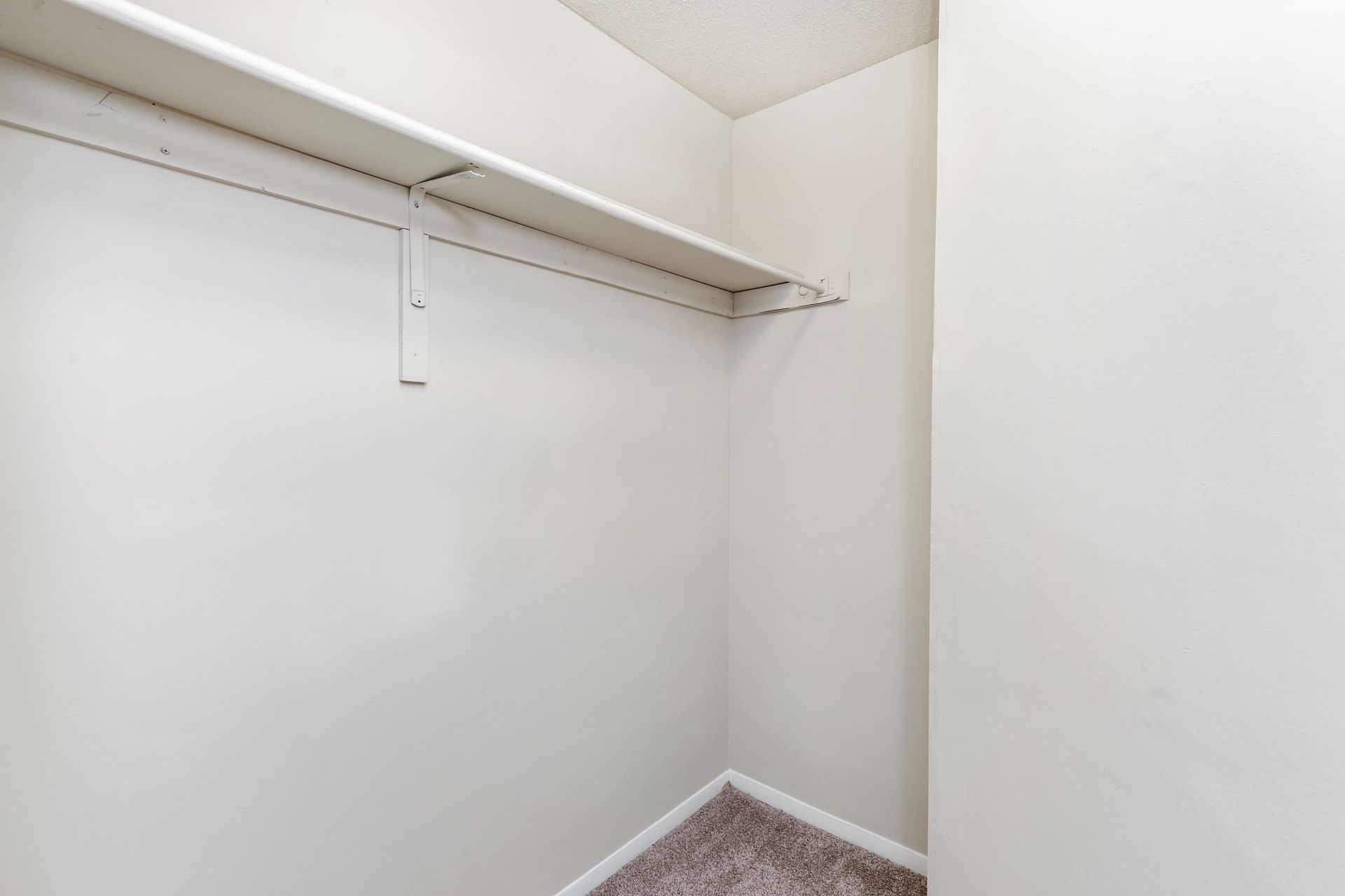 An empty, neutral-toned walk-in closet featuring a single white wire shelf mounted on a wall above carpeted flooring.