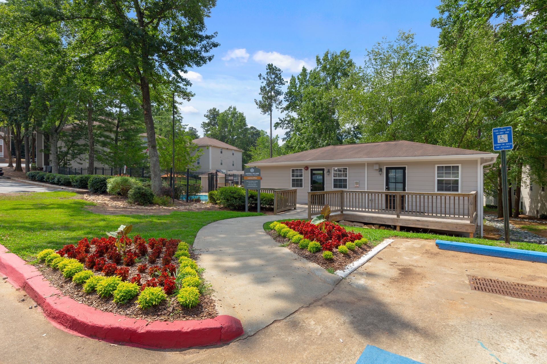 A paved path leads to a low-rise apartment building with a wooden ramp, surrounded by lush trees and manicured gardens.