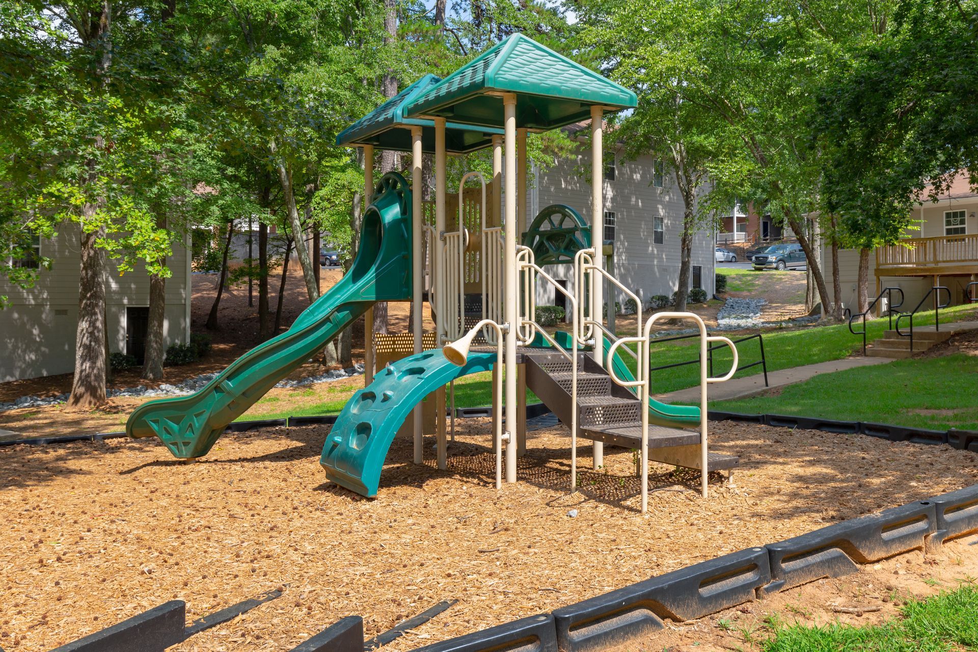 A playground structure with a green roof, a slide, climbing wall, and stairs, set in a woodchip area surrounded by trees.