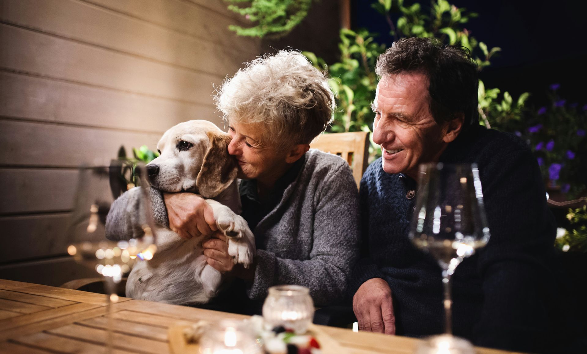 Two people smiling at a table, holding a small white dog, with wine glasses and warm evening lights.