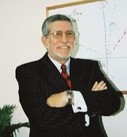 Man in a black suit with red tie, arms crossed, standing before a whiteboard with charts