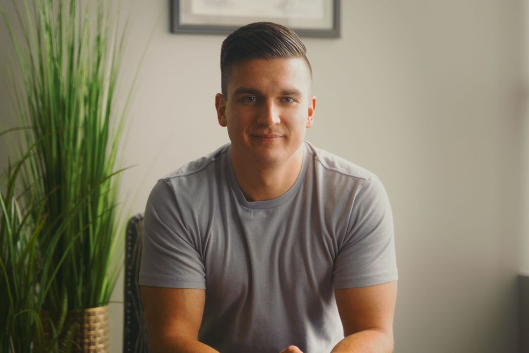 Person in a gray shirt sitting indoors beside a tall green plant, with a framed picture on the wall.