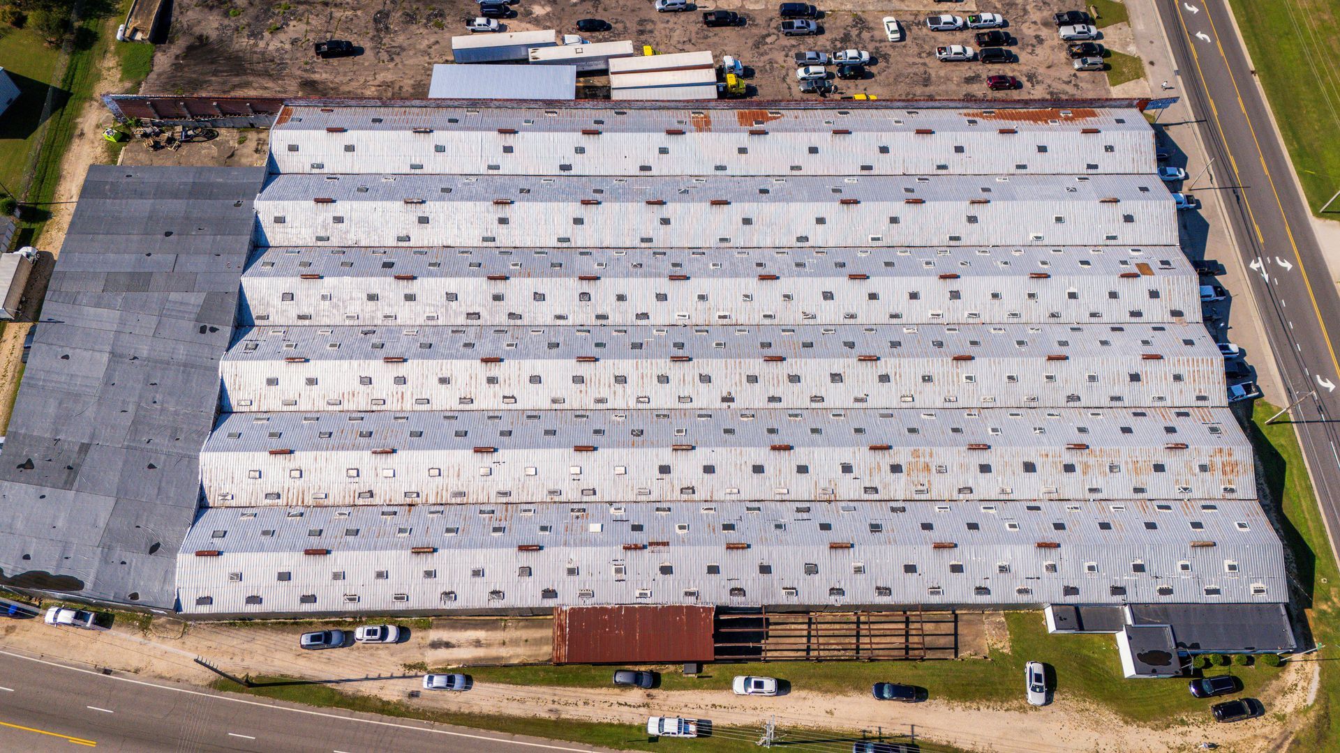 Aerial view of a large white-roofed industrial warehouse with trucks and parked cars around it.