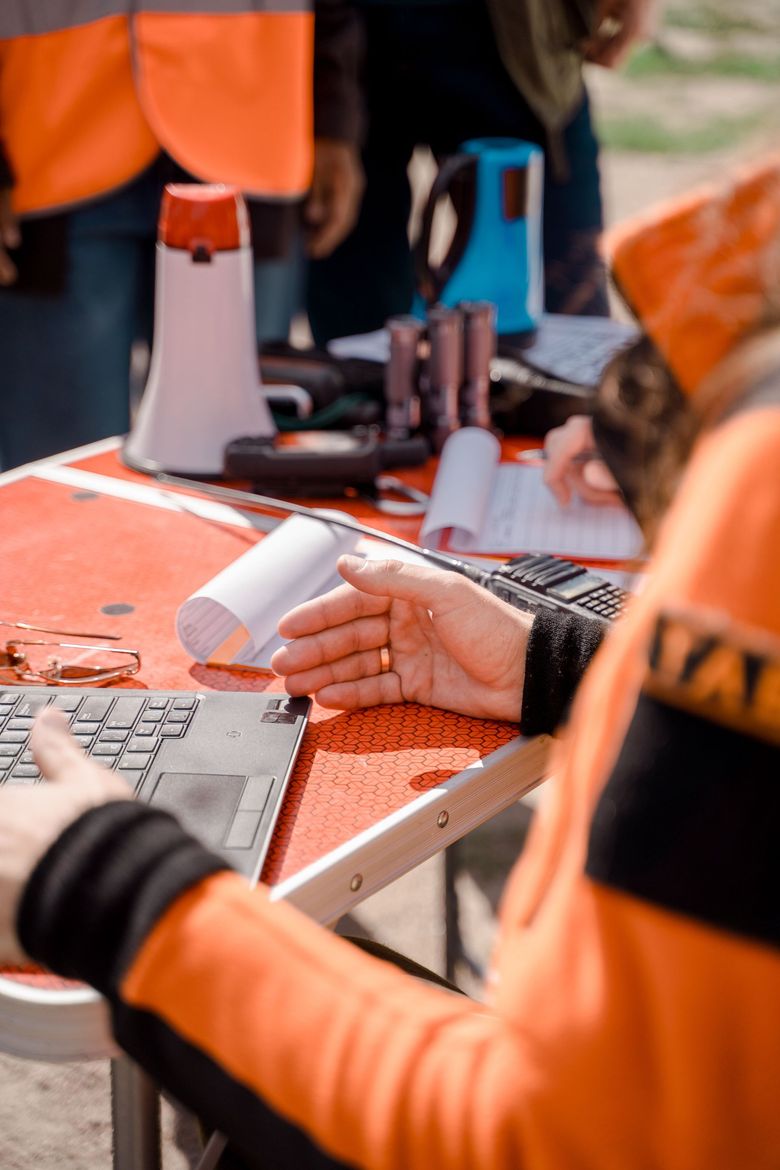 People in orange safety vests using tools and papers at an outdoor worktable