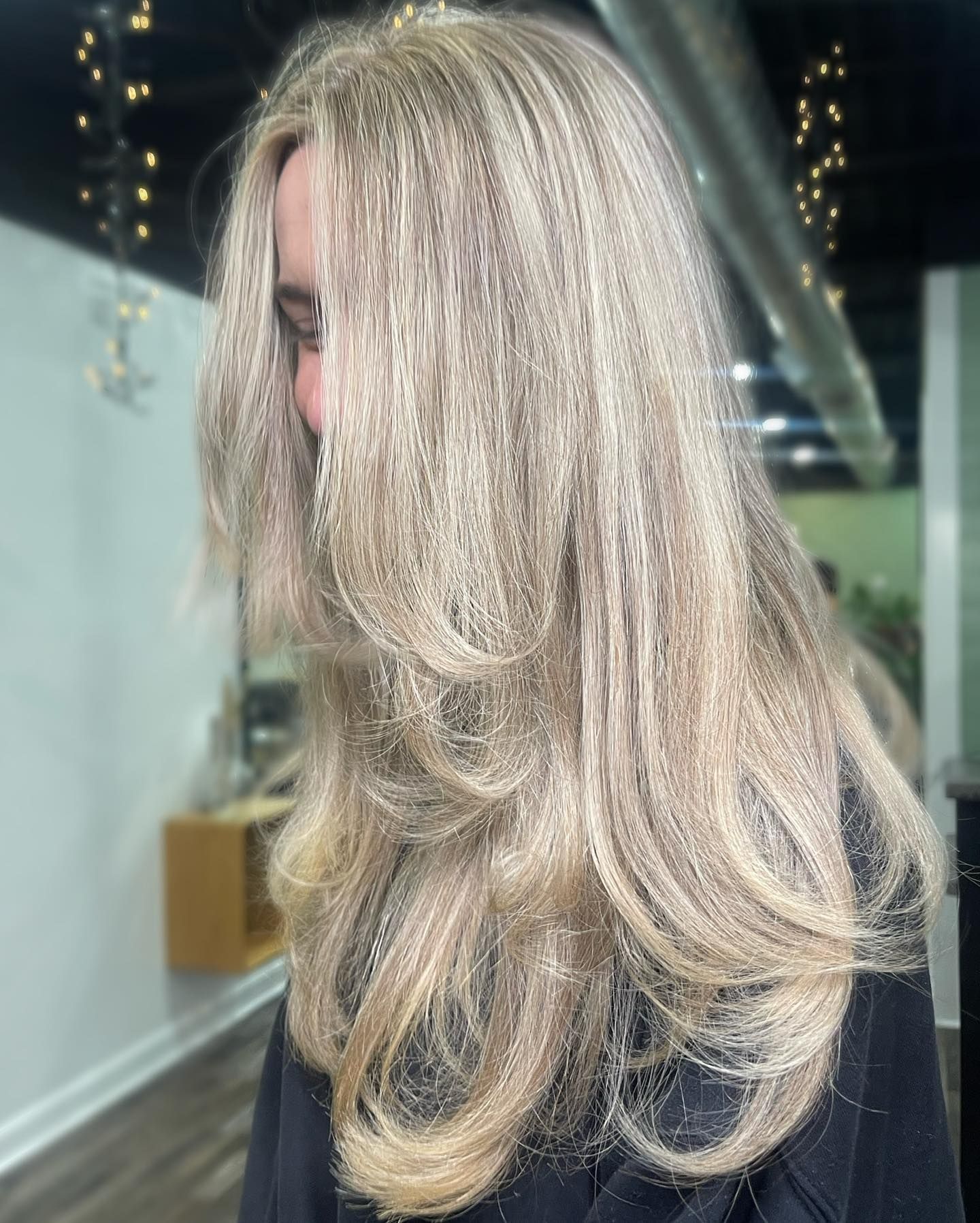 Woman with long, layered, light blonde hair. Indoors, blurred background, possibly a salon.