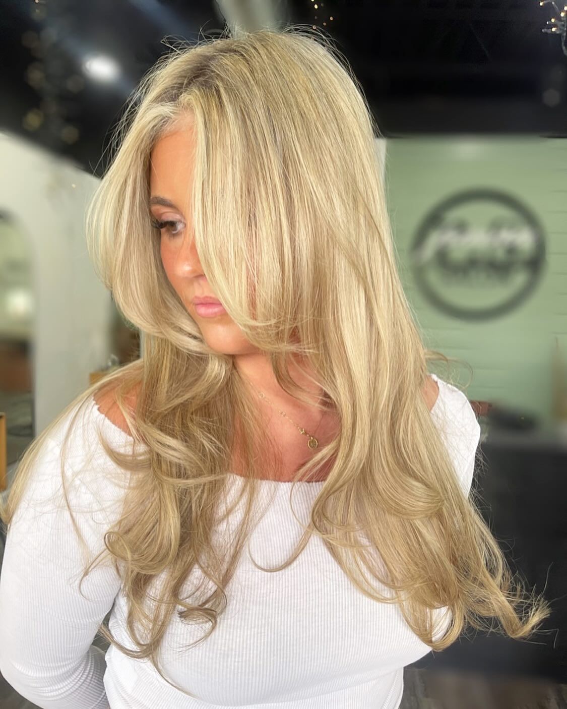 Blonde hair styled with crimped top layer, face partially obscured, wearing a white off-shoulder shirt.
