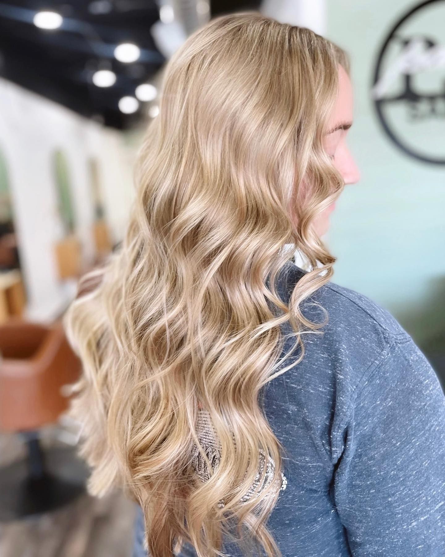 Woman with long wavy blonde hair in a salon, wearing a dark blue shirt.