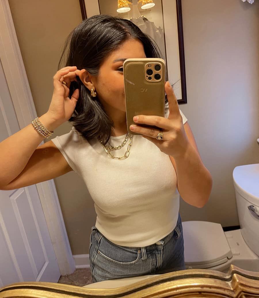 Woman in a bathroom taking a mirror selfie, wearing a white top and holding her hair.