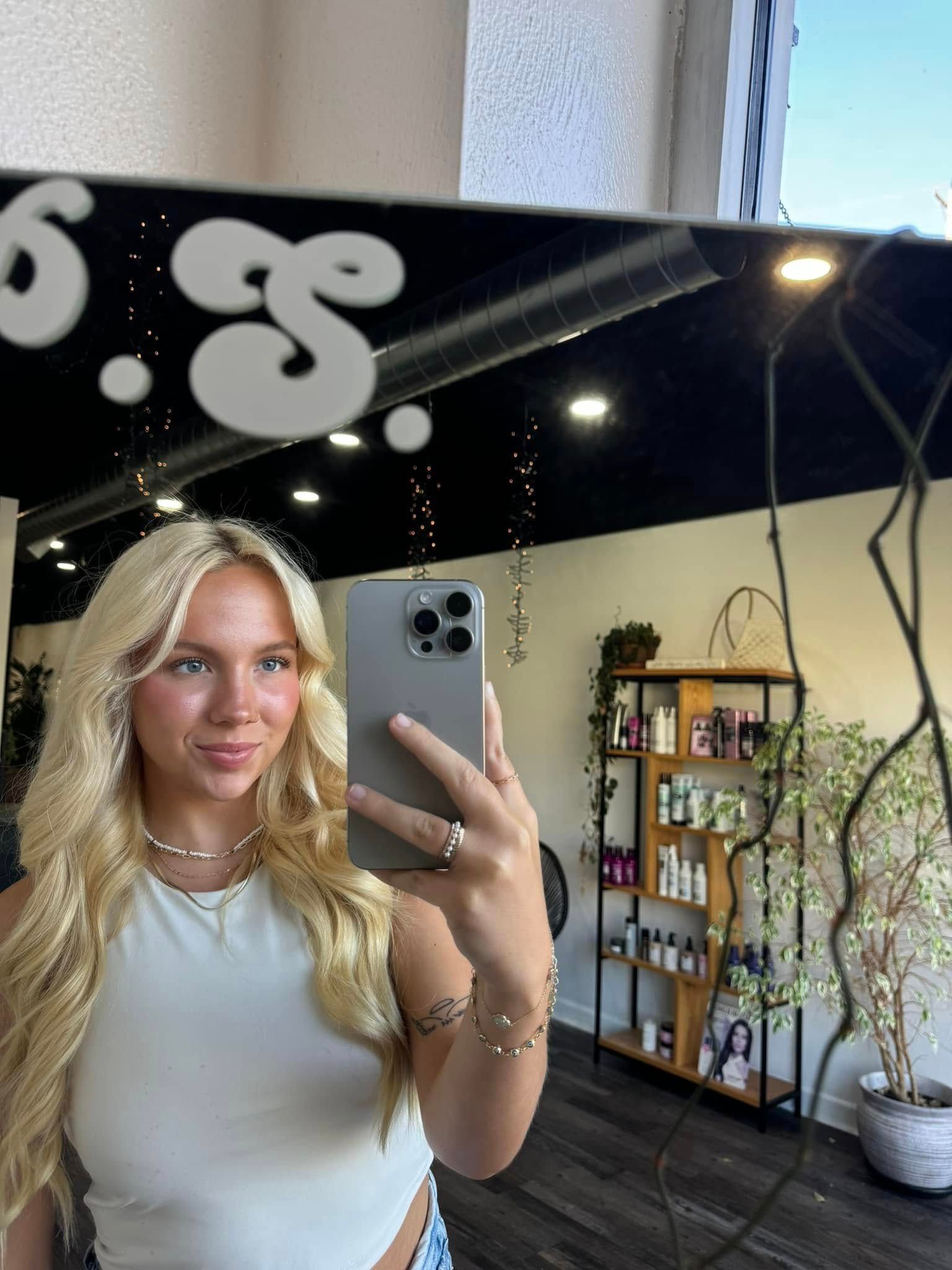 Blonde woman takes selfie in salon mirror. She wears a white top and jewelry, smiling.