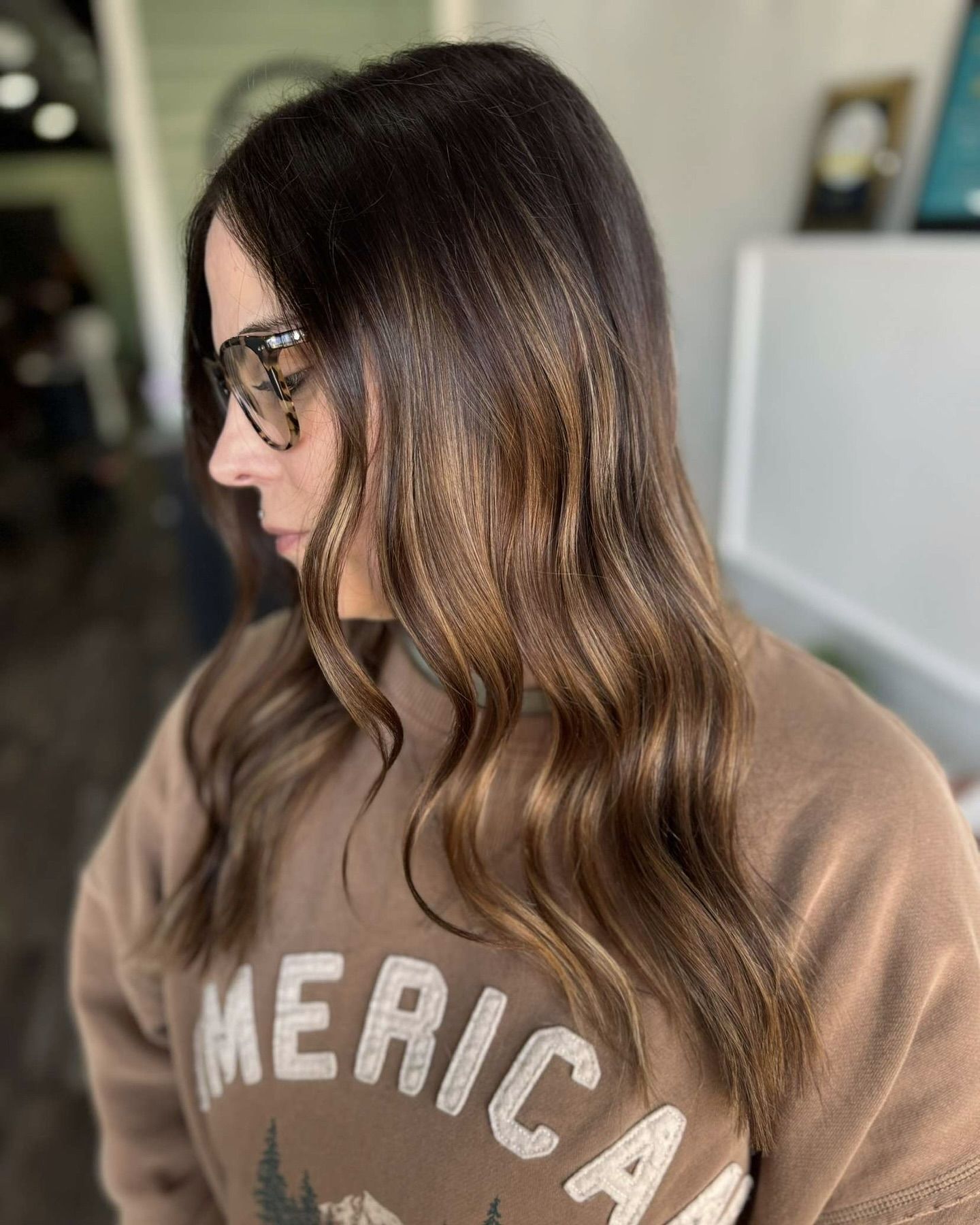 Woman with dark brown hair with highlights, wearing glasses and a brown sweatshirt with 