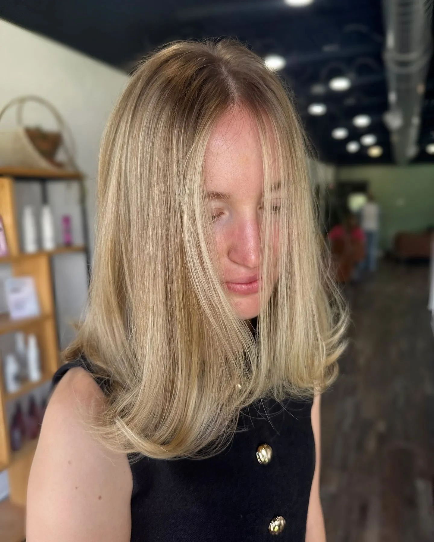 Person with shoulder-length blonde hair in a black top, standing indoors in a softly lit room.