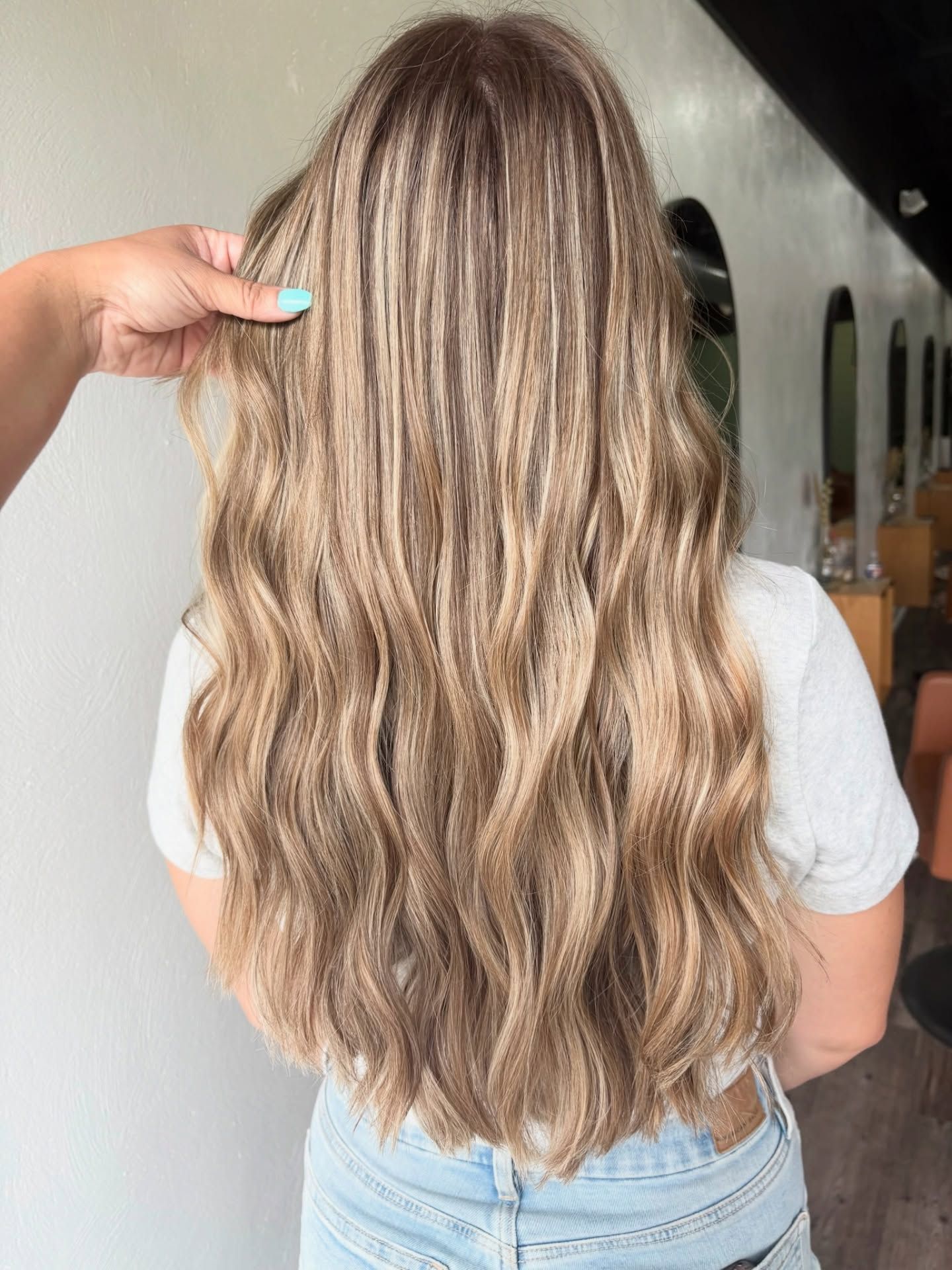 Woman with long, wavy blonde hair with highlights, standing in a salon. A hand touches the hair.