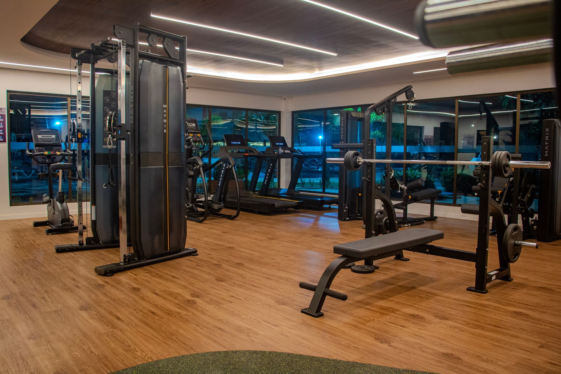 Academia moderna com aparelhos de musculação, bancos e grandes janelas com vista para a cidade à noite.