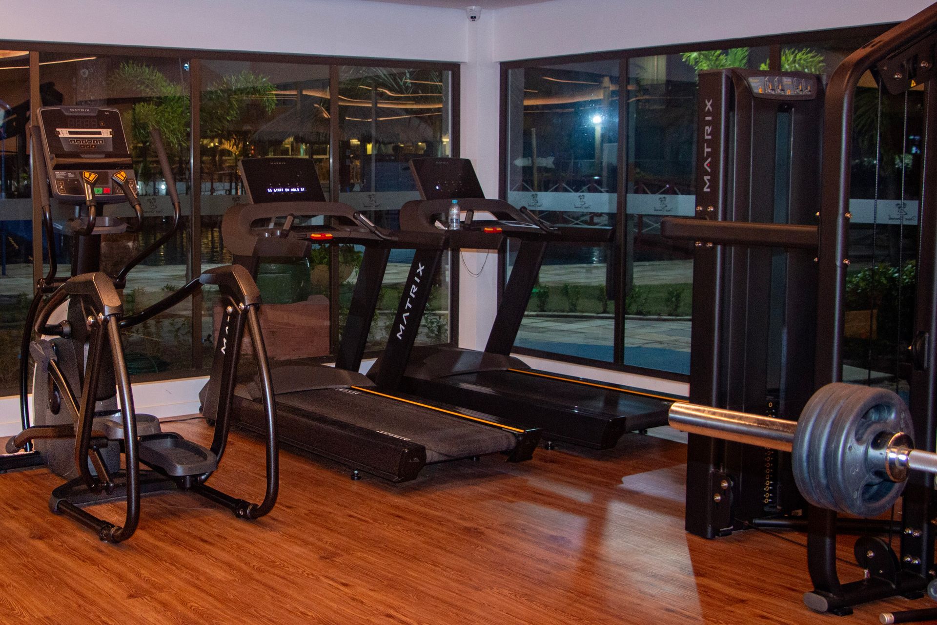 Academia com aparelhos de cardio, pesos e grandes janelas com vista para a vegetação à noite.